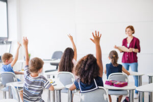 Rear view of school children sitting at the desk in classroom, raising hands.