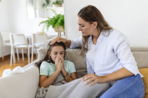 Young woman checking temperature with hand of little ill daughter. Mother checking temperature of her sick girl. Sick child lying on bed under blanket with woman checking fever on forehead by hand.